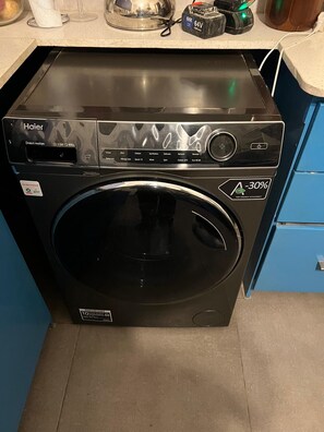 Vista frontal de una lavadora Haier con puerta de carga frontal, panel de control con múltiples opciones y una etiqueta que indica máxima eficiencia energética.