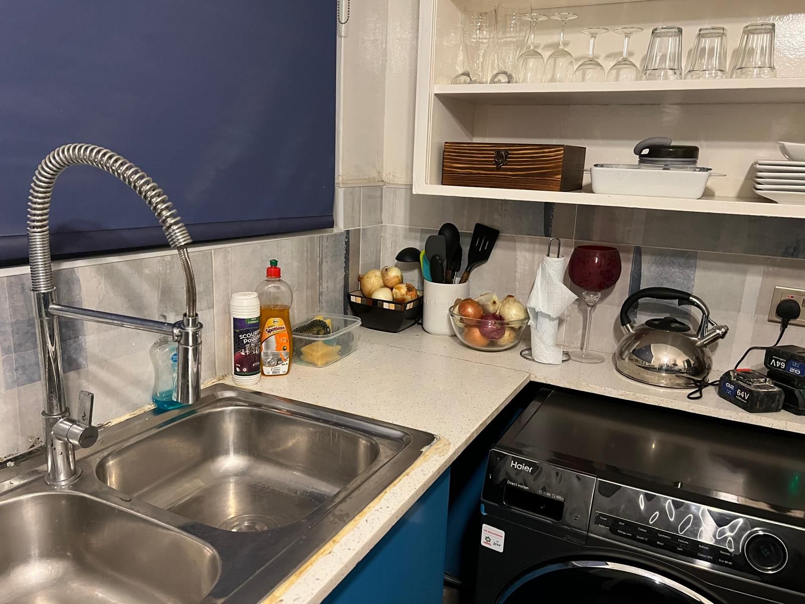 Ett kök med diskhoar i rostfritt stål, ett diskställ, en vattenkokare och en tvättmaskin.