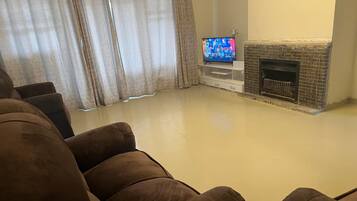 Ein Wohnzimmer mit Flachbildfernseher auf einem Standfuß, Kamin und einer großen braunen Couch.