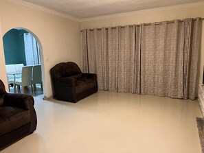 A spacious living room with a dark brown sofa, a dining table with chairs, and a large window with curtains.
