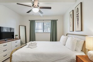 Un dormitorio con cama, televisor, ventilador de techo, ventana con cortinas y cuadros en la pared.