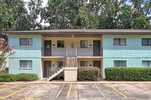 A two-story building with a balcony on the second floor, a parking lot in front, and a hedge to the right. - 2BR Savannah condo w/ pool, WiFi, parking, full kitchen * 30+ days (Savannah)