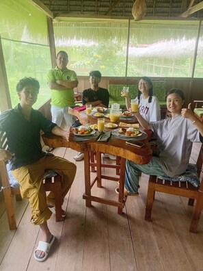 Un groupe de personnes est assis autour d’une table en bois, devant de la nourriture et des boissons, dans une pièce dotée d’un toit de chaume et de grandes fenêtres.