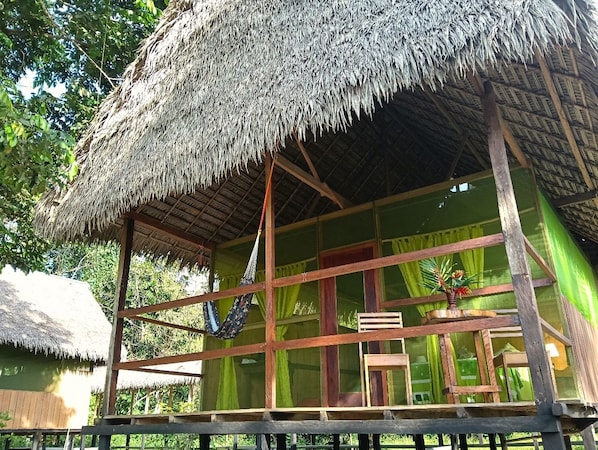 A thatched-roof hut with a wooden frame, a hammock, and a window with curtains.