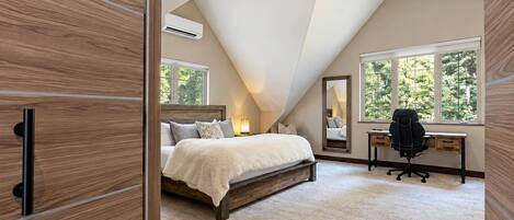 A bedroom with a sloped ceiling, a bed, a desk, and a large window.
