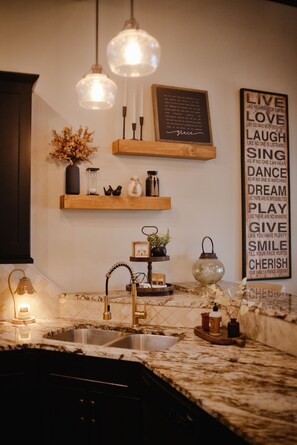 A kitchen with a marble countertop, a sink, pendant lights, and decorative items on shelves. - Lakefront Retreat on Greers Ferry Lake with EV Charger (Higden)