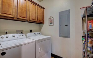 A laundry room with two white Whirlpool washers and dryers, wooden cabinets above, a small framed inspirational quote on the wall, and a metal shelf with cleaning supplies. - Lakefront Retreat on Greers Ferry Lake with EV Charger (Higden)