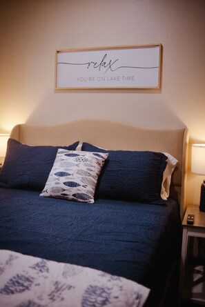 A bedroom with a bed, pillows, a bedside table with a lamp, and a wall with a framed sign.