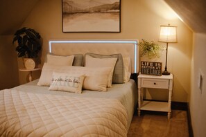 A neatly made bed with a quilted cover and multiple pillows, a bedside lamp, a small bedside table with a plant, and a framed picture on the wall.