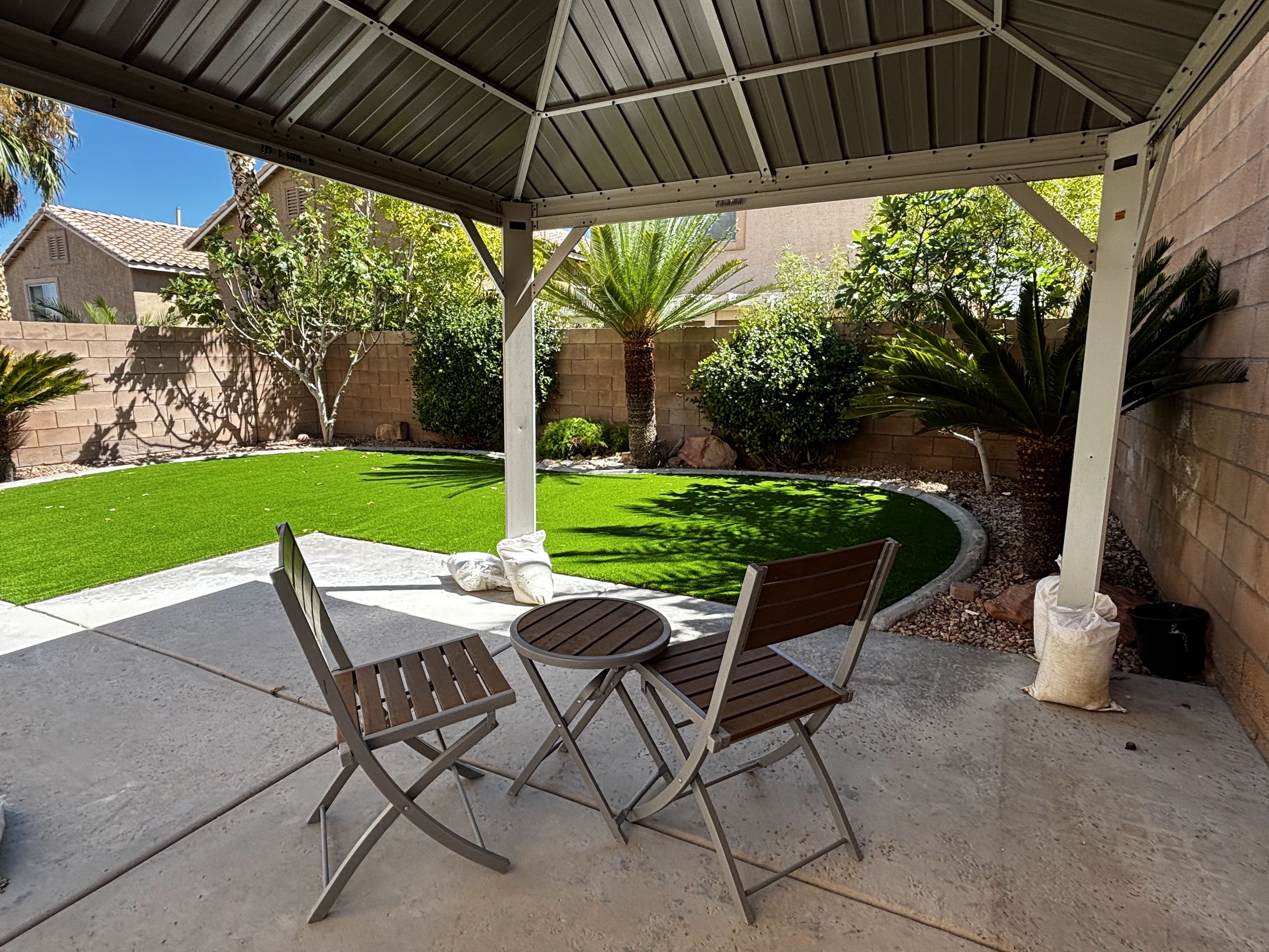 A covered patio with two wooden chairs and a table, surrounded by a well-maintained lawn and various plants.