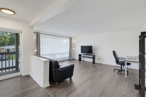 A modern living room with a black leather sofa, a flat-screen TV, a desk with a chair, and a large window with curtains.