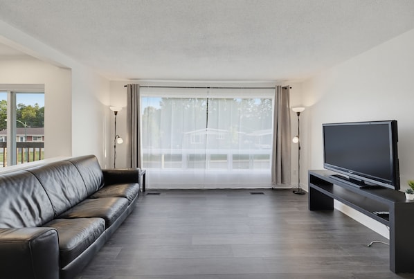 A modern living room with a leather sofa, a flat-screen TV on a stand, and a large sliding glass door leading to a balcony.