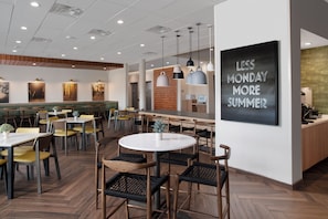 A modern restaurant interior with wooden flooring, round tables, and chairs. There are pendant lights and a chalkboard with 'LESS MONDAY MORE SUMMER' written on it. - Fairfield by Marriott Inn & Suites Cedar Park (Leander)