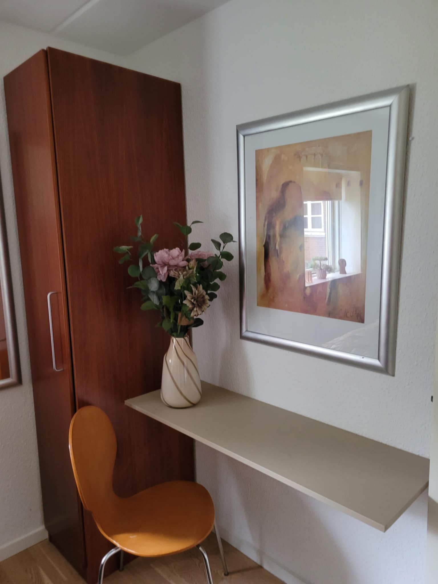 Une pièce comprenant une armoire en bois, une chaise, un vase avec des fleurs et un tableau abstrait encadré accroché au mur.