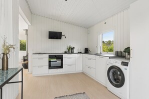 A modern kitchen with white cabinetry, a built-in oven, a washing machine, and a flat-screen TV mounted on the wall. - Cozy holiday home in Klint – Nykøbing Sj. (Nykøbing Sjælland)