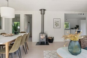 A modern interior with a central wood-burning stove, a dining area with chairs and a table, and a kitchen in the background. - Cozy holiday home in Klint – Nykøbing Sj. (Nykøbing Sjælland)