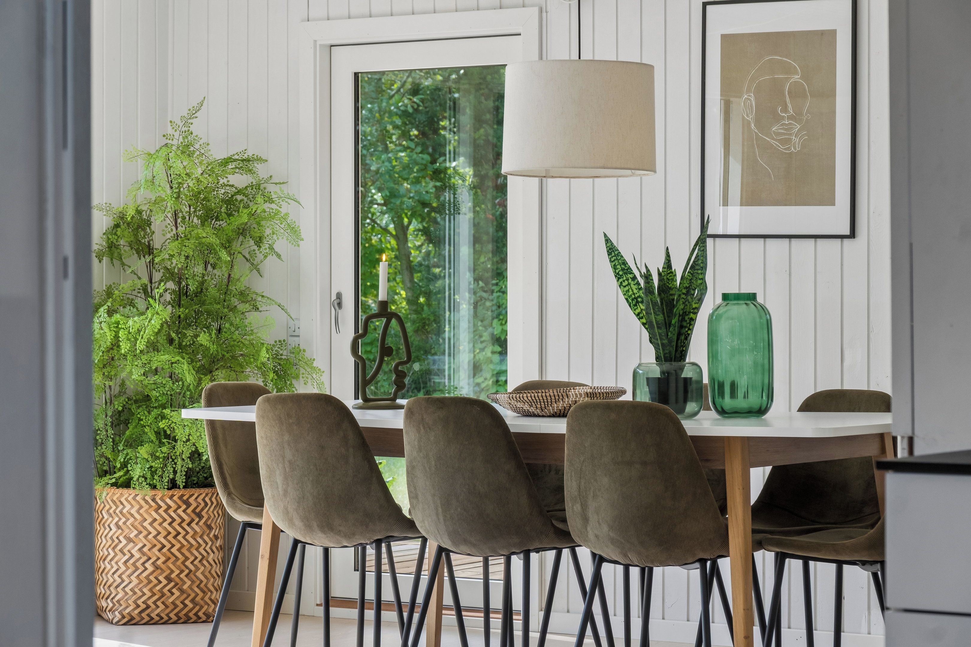 Un comedor con una mesa blanca, sillas marrones, una planta verde y un cuadro en la pared.