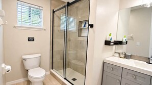 Ein Badezimmer mit Toilette, einer Duschkabine, einem Waschbecken mit Spiegel und einem Fenster mit Jalousien.