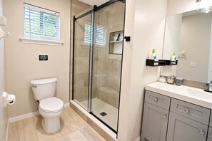Een badkamer met toilet, een douchecabine, een wastafel met spiegel en een raam met jaloezieën.