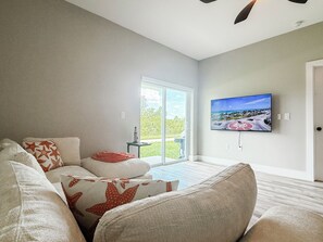 A modern living room with a large flat-screen TV mounted on the wall, a sliding glass door leading to a balcony, and a sectional sofa with patterned cushions. - 20 Min to Englewood Beach • Park + BBQ (Port Charlotte)