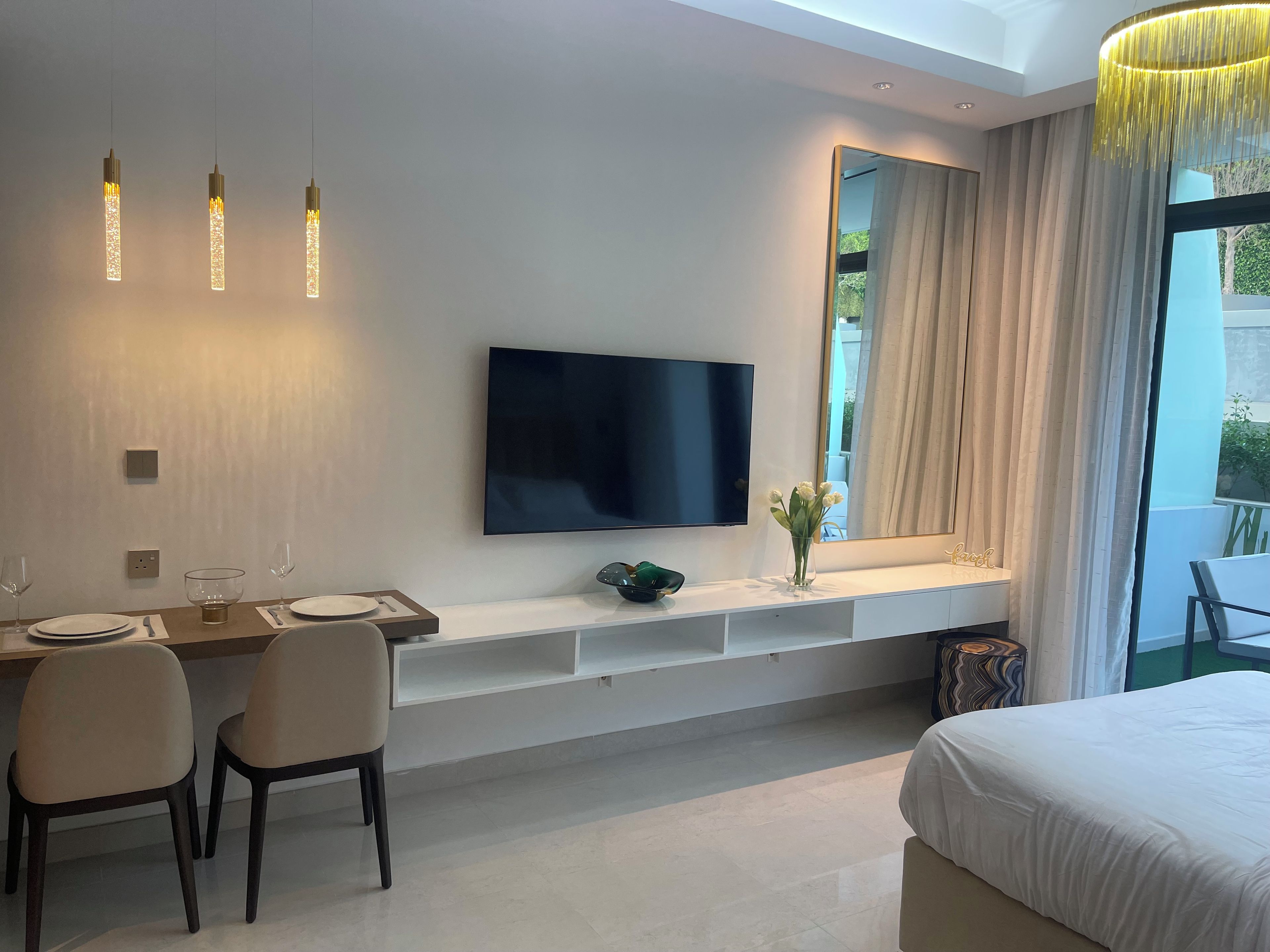 A modern hotel room with a bed, a dining area with chairs and a table, a TV mounted on the wall, and a large window with curtains.
