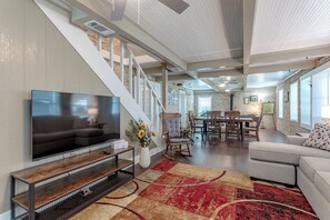 A modern living room with a flat-screen TV, a wooden entertainment unit, a red and beige rug, a grey sofa, and a dining area with a table and chairs. - Historic 1800s Rock House w/ horse corral (Gatesville)