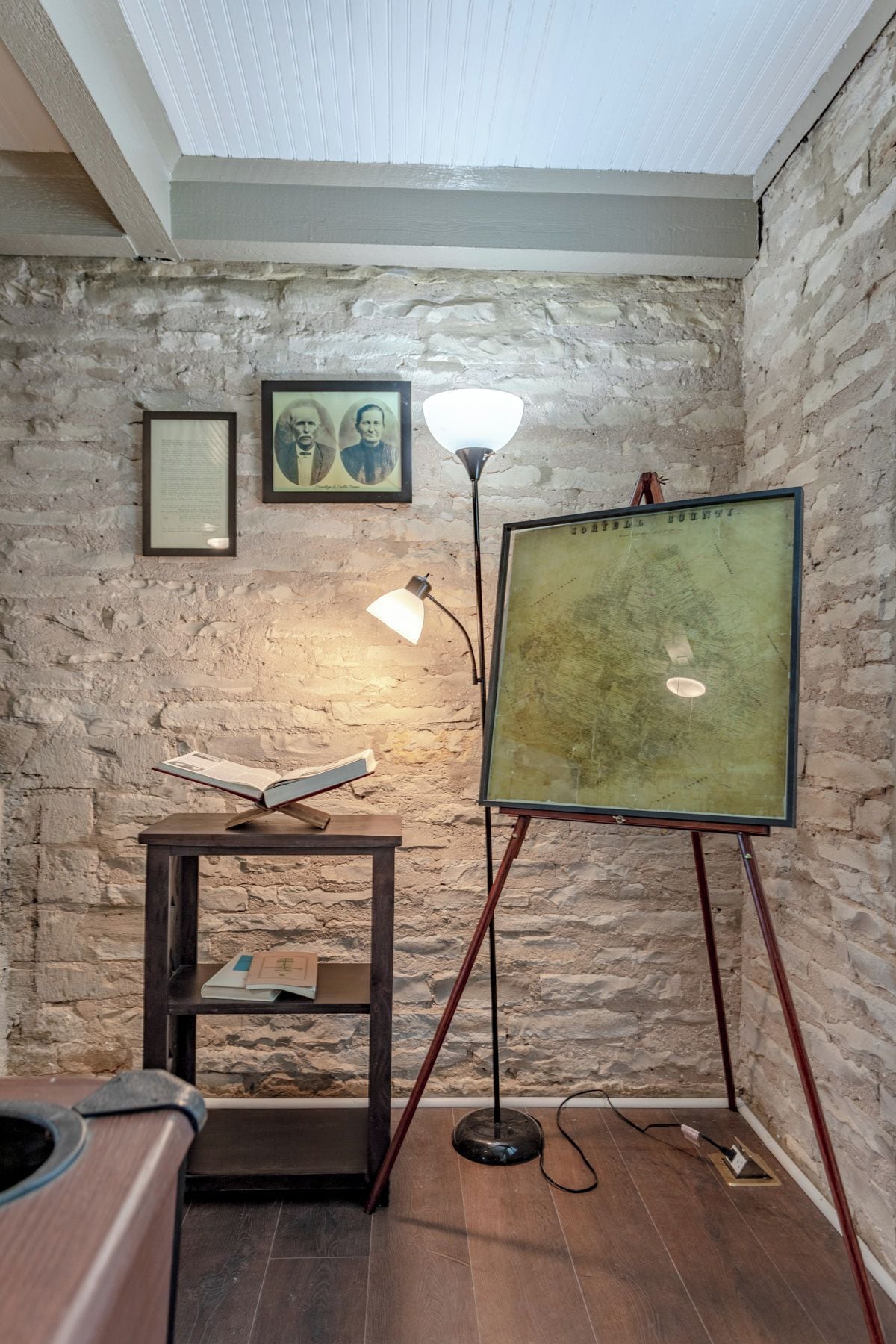 A room with a stone wall, a wooden table with a book, a floor lamp, and a framed picture on the wall.