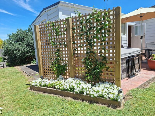 A wooden lattice structure with climbing plants and white flowers, a patio area with a table and chairs, and a white building in the background.