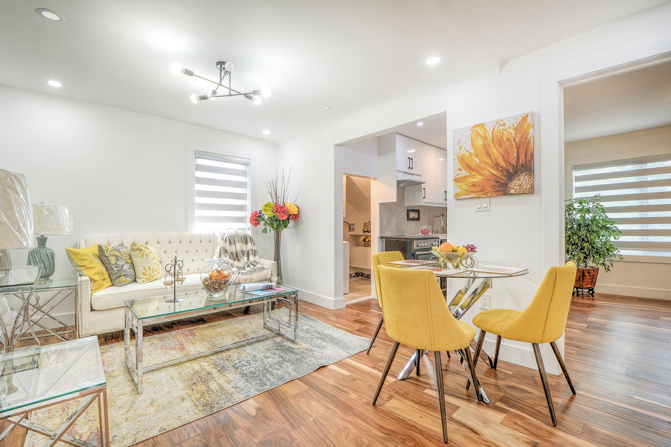 A modern living room with a glass coffee table, a sofa with cushions, and a dining area with yellow chairs.