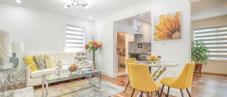 A modern living room with a glass coffee table, a sofa with cushions, and a dining area with yellow chairs.