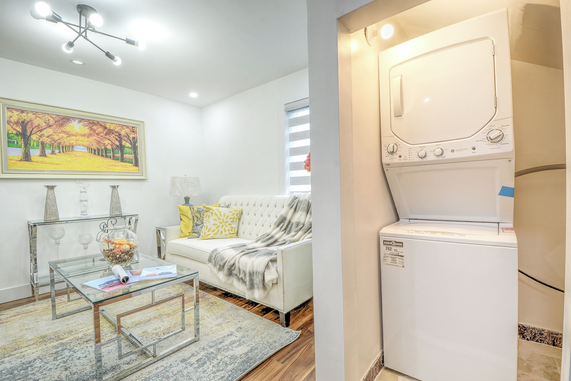 A living room with a sofa, a glass coffee table, a painting, and a white washing machine.