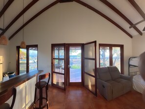 A spacious room with a high ceiling, a dining area, a seating area with a sofa, and large windows offering a view of the outdoors.