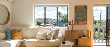 Uma sala de estar moderna com um sofá branco, um espelho redondo, uma pequena mesa redonda e vista para o exterior.
