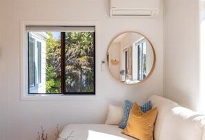 A room with a white couch, a round mirror, and a window with a view of greenery.