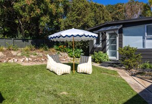 A backyard with a lawn, two striped chairs, a white umbrella, and a house with a window.