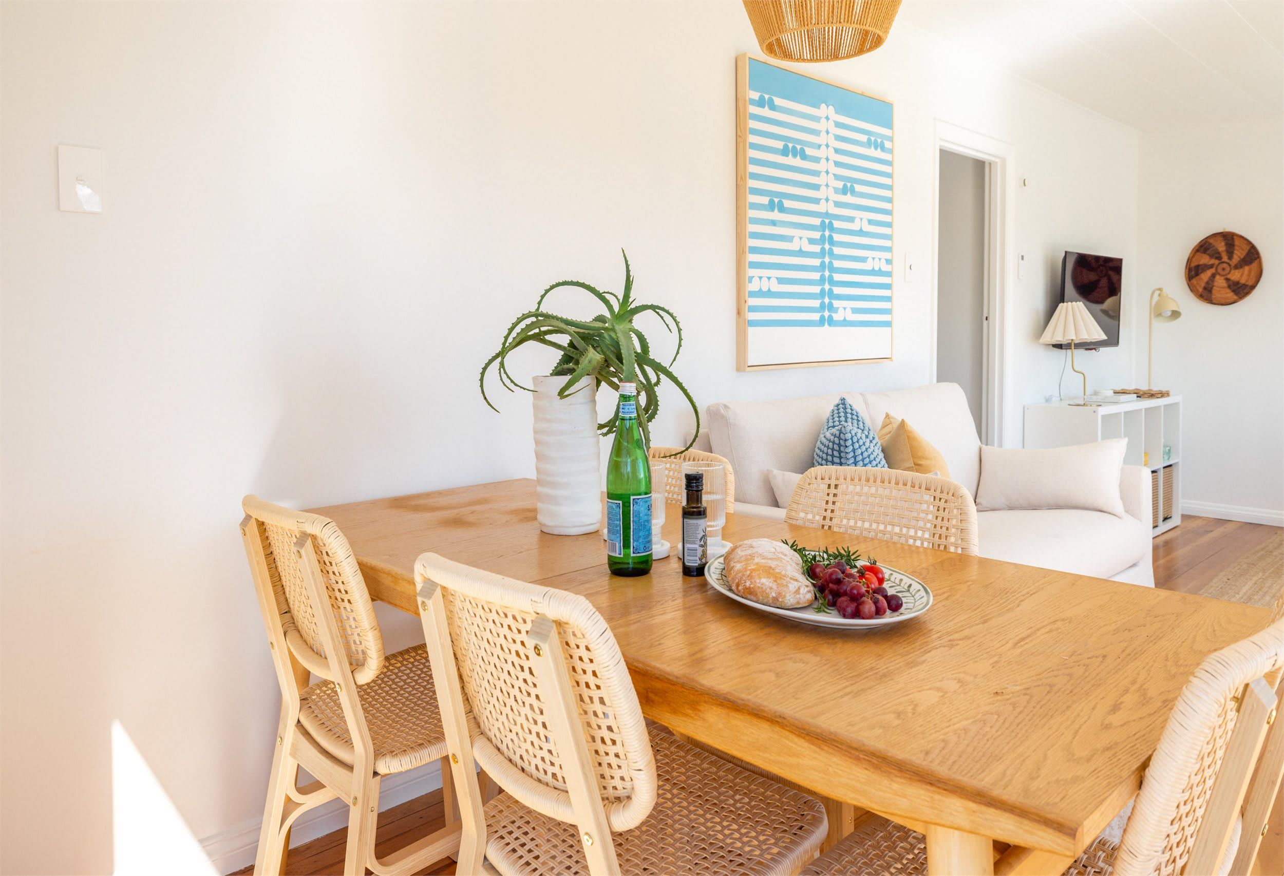 A dining area with a wooden table, wicker chairs, a white sofa, and a wall-mounted painting.