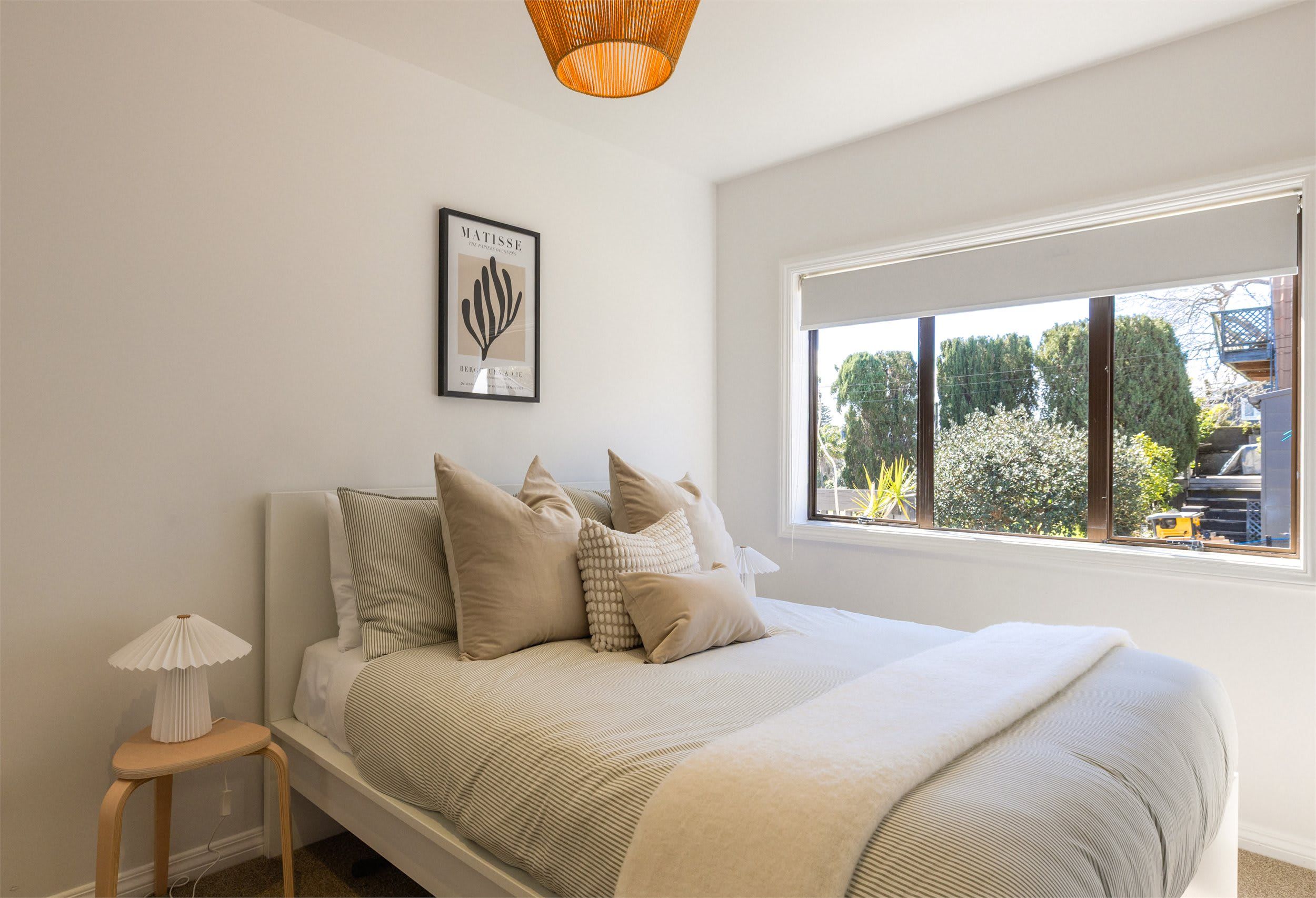 A bedroom with a bed, pillows, a bedside table, a lamp, a window with a view, and a wall-mounted artwork.