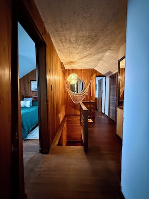 A wooden interior with a hammock, a staircase, and a bed with a blue bedspread. - Charming 3-bedroom Cottage in Cleverdale on Beautiful Lake George with AC, WiFi (Queensbury)