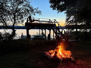 A wooden deck overlooking a lake with a fire pit in the foreground. - Charming 3-bedroom Cottage in Cleverdale on Beautiful Lake George with AC, WiFi (Queensbury)