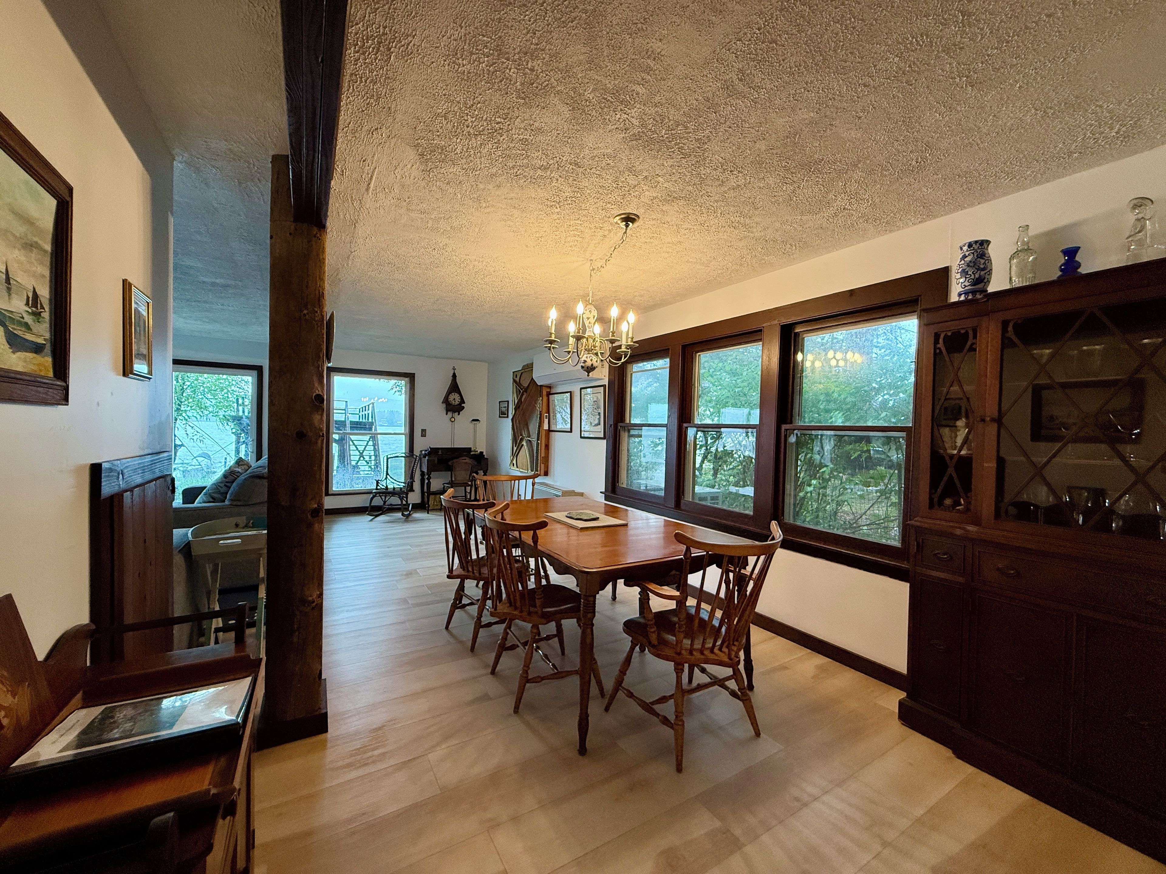 A dining area with a wooden table and chairs, a chandelier, and large windows with a view of greenery.