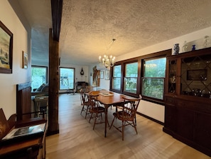 A dining area with a wooden table and chairs, a chandelier, and large windows with a view of greenery. - Charming 3-bedroom Cottage in Cleverdale on Beautiful Lake George with AC, WiFi (Queensbury)