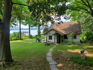 A house with a stone pathway leading to the front door, surrounded by lush greenery and a view of a lake. - Charming 3-bedroom Cottage in Cleverdale on Beautiful Lake George with AC, WiFi (Queensbury)
