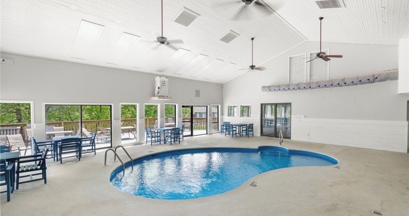 A large indoor swimming pool with ceiling fans, a sunroom with large windows, and outdoor seating.