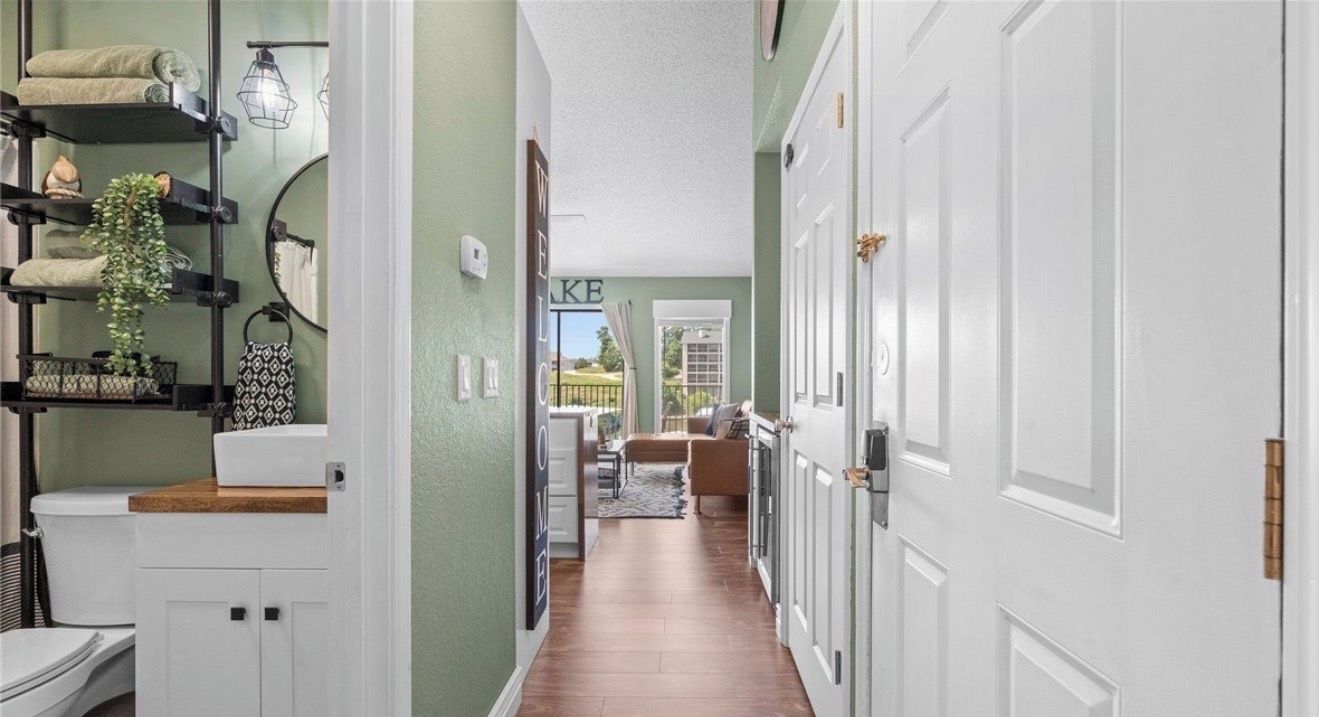 Un pasillo con una puerta blanca, un letrero de bienvenida y vista a una sala de estar con alfombra y muebles.