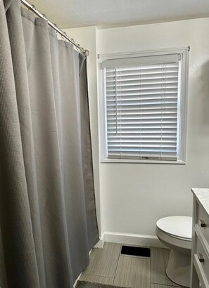 A bathroom with a shower curtain, a window with blinds, a toilet, and a sink.