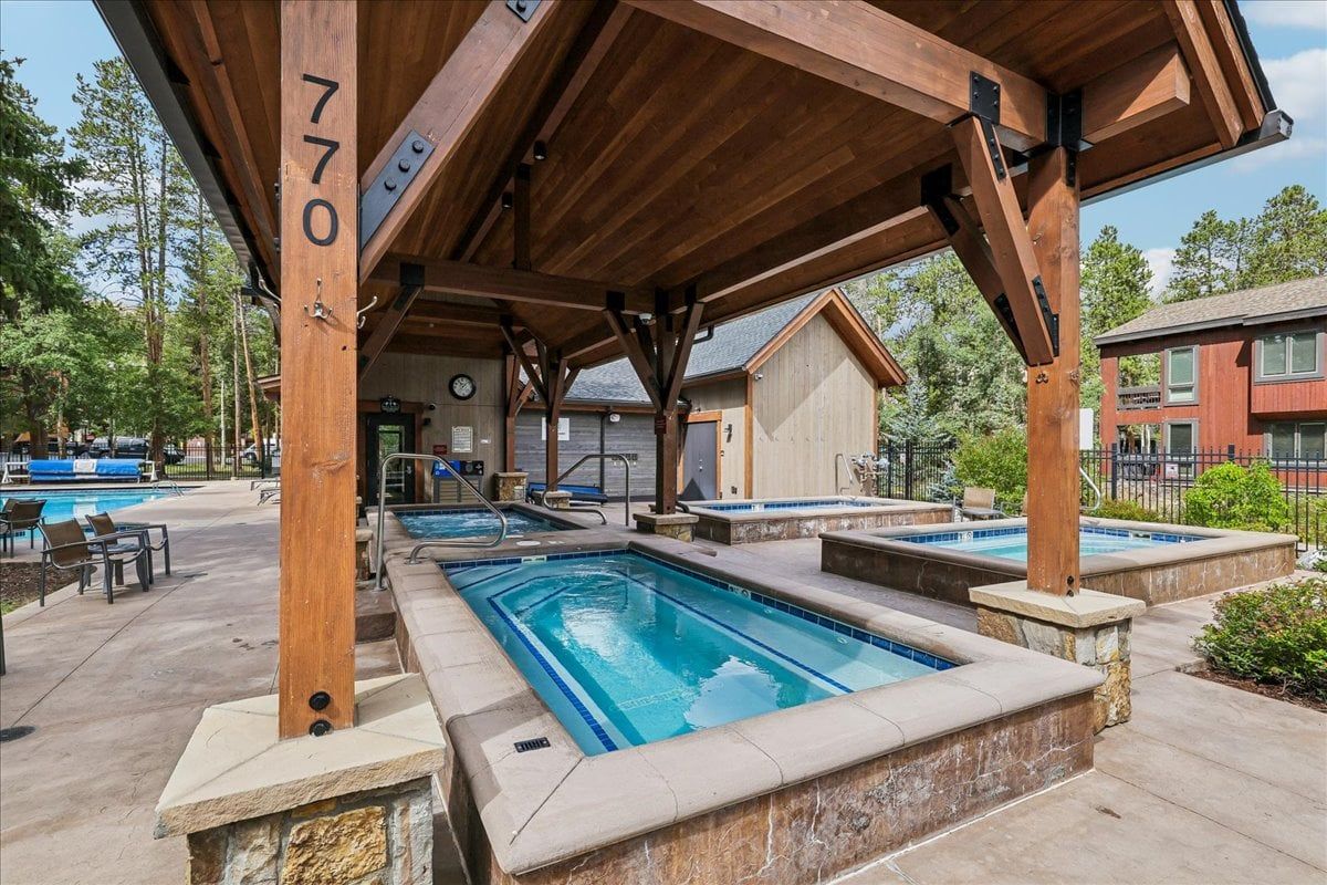 A pool area with multiple pools under a wooden structure, a clock, and a building in the background.