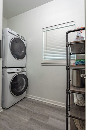 A laundry room with two stacked front-loading washers and dryers, a window with blinds, a metal shelf with cleaning supplies, and a wooden floor. - Bear Basecamp/3BR/Game Room/Midterm Welcome (Waco)