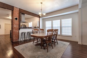 A dining area with a wooden table and chairs, a kitchen in the background, and large windows with blinds. - Bear Basecamp/3BR/Game Room/Midterm Welcome (Waco)