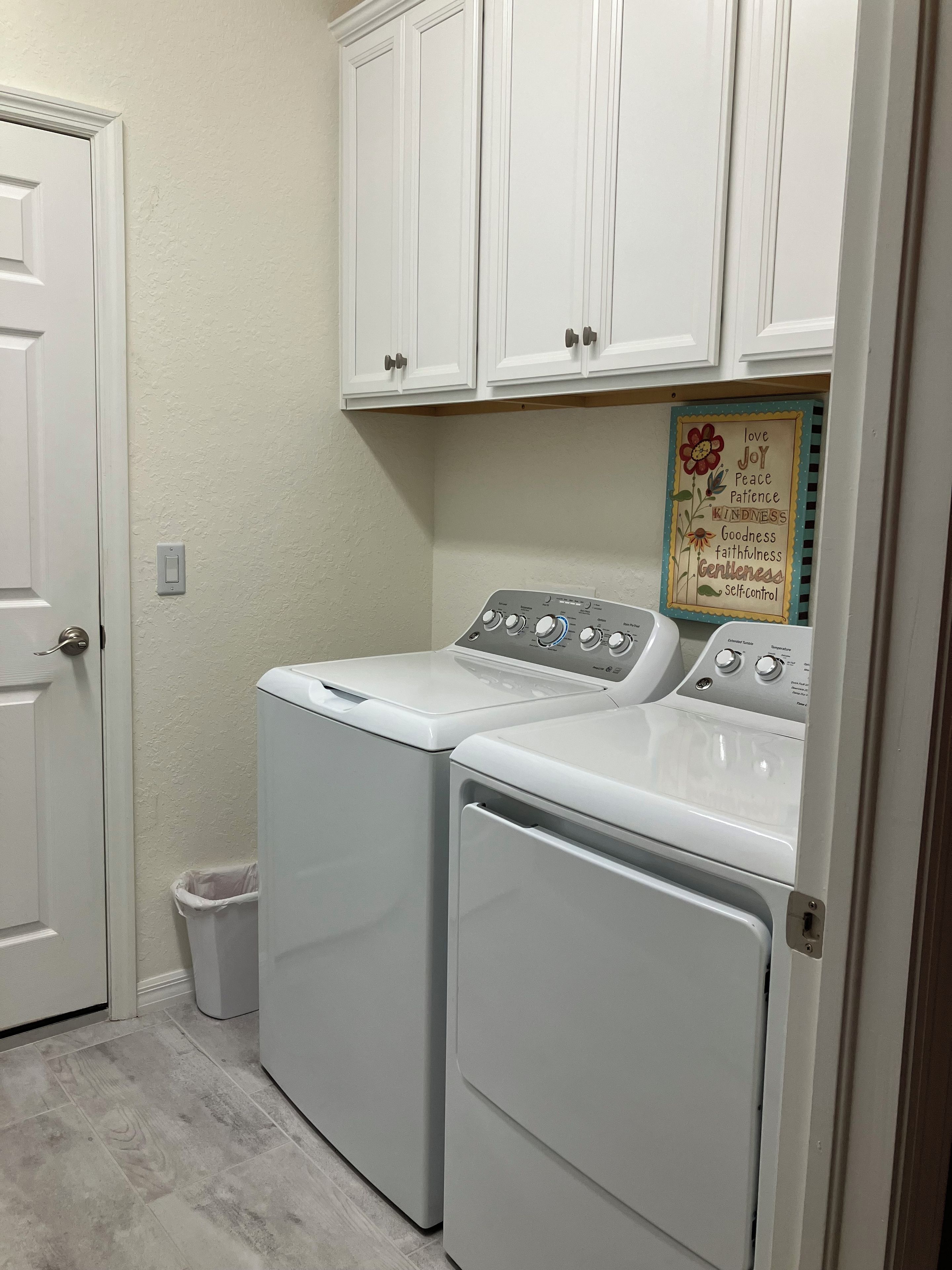 A laundry room with two front-loading washing machines, a white door, a small trash bin, and a wall-mounted cabinet above the machines.