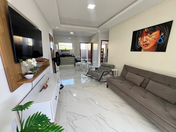 A modern living room with a large flat-screen TV, a sofa, a dining area, and a kitchen in the background.